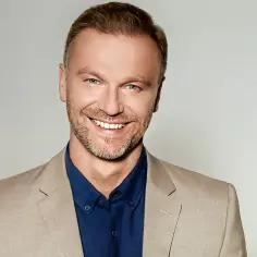 Uśmiechnięty Krystian Wieczorek w beżowej marynarce i granatowej koszuli, gotowy do kolejnych filmów.