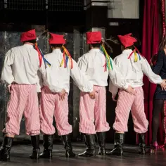 Czterech mężczyzn w strojach ludowych, z kolorowymi wstążkami przy czapkach, stoi tyłem do widza. Obok nich kobieta w błyszczącej sukience. Scena z przedstawienia "goło i wesoło spektakl".