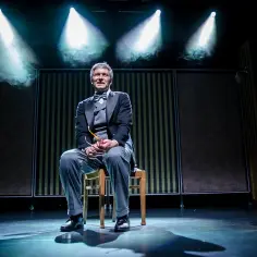 Aktor w garniturze siedzi na krześle na scenie, obok niego dziecko w okularach.