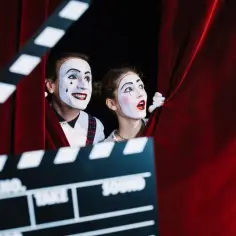 Dwoje mimów z makijażem, zasłoniętych czerwoną kurtyną, z klapsem filmowym na pierwszym planie. Marzą, jak zostać aktorką.
