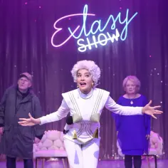 Voucher do teatru dla dwojga Warszawa: aktorka w białym stroju z frędzlami, mężczyzna w zielonej kurtce i kobieta w niebieskiej sukience na scenie.