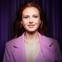 Anna Karczmarczyk w różowej marynarce, z rudymi włosami i niebieskimi oczami, uśmiecha się na tle fioletowej kurtyny. Jej filmy, seriale i programy to gwarancja dobrej rozrywki.