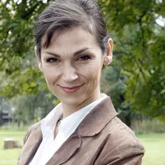 Olga Bończyk w brązowej marynarce i białej koszuli, uśmiecha się na tle drzew.