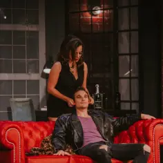 Scena z najlepszych komedii teatralnych: kobieta w czarnej sukience pochyla się nad mężczyzną w skórzanej kurtce, siedzącym na czerwonej kanapie.