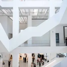 Wnętrze muzeum sztuki nowoczesnej z białymi, geometrycznymi schodami i balustradami. Ludzie zwiedzają, kupując bilety i podziwiając ekspozycje.