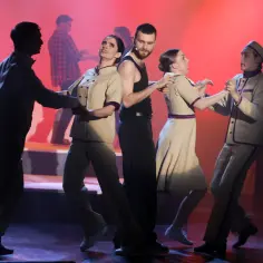 Aktorzy w strojach z epoki tańczą na scenie z czerwonym tłem.