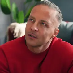 Justin z "Barw Szczęścia" w czerwonym swetrze, zamyślony, w tle kanapa z kolorowymi poduszkami.