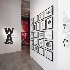 Wystawa w muzeum sztuki nowoczesnej prezentuje prace Maryny Tomaszewskiej. Na ścianie widnieje logo WA oraz grafiki z datami i tytułami.