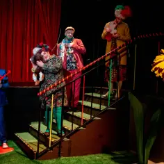 Aktorzy w barwnych strojach i perukach podczas sztuki teatralnej po angielsku. Na scenie wielka żółta kwiatowa dekoracja.