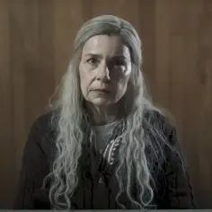 Agata Kulesza w serialach i programach, z długimi, siwymi włosami i poważnym wyrazem twarzy, siedzi przed drewnianą ścianą.