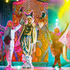 Kolorowy teatr dla nastolatków: aktorzy w fantazyjnych strojach, w tym postać z kocimi uszami, tańczą na scenie.