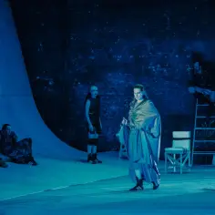 Scena teatralna w niebieskim świetle. Grupa aktorów w nietypowych strojach, być może spektakl z repertuaru teatrów Warszawa dziś.