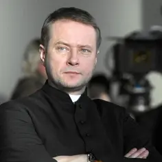 Artur Żmijewski w roli księdza, jak w wielu jego filmach. Skupiony, z rękami skrzyżowanymi na piersi, na planie filmowym.
