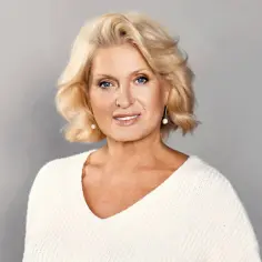 Uśmiechnięta Ewa Kasprzyk w białym swetrze. Jej blond włosy i niebieskie oczy przyciągają uwagę, jak w jej najlepszych filmach.
