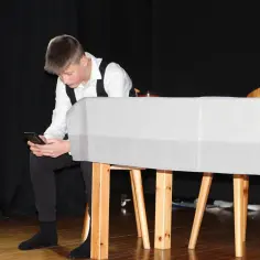Chłopiec w białej koszuli i kamizelce, co robi scenograf, sprawdza coś na telefonie, siedząc przy scenografii.