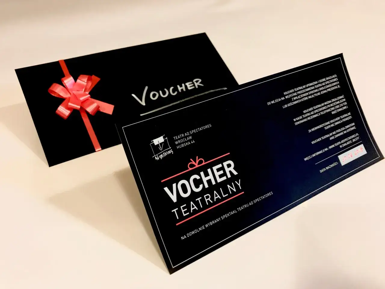 Czarny voucher teatralny z czerwoną kokardką. Idealny prezent, by wybrać się na spektakl do teatru w Warszawie.