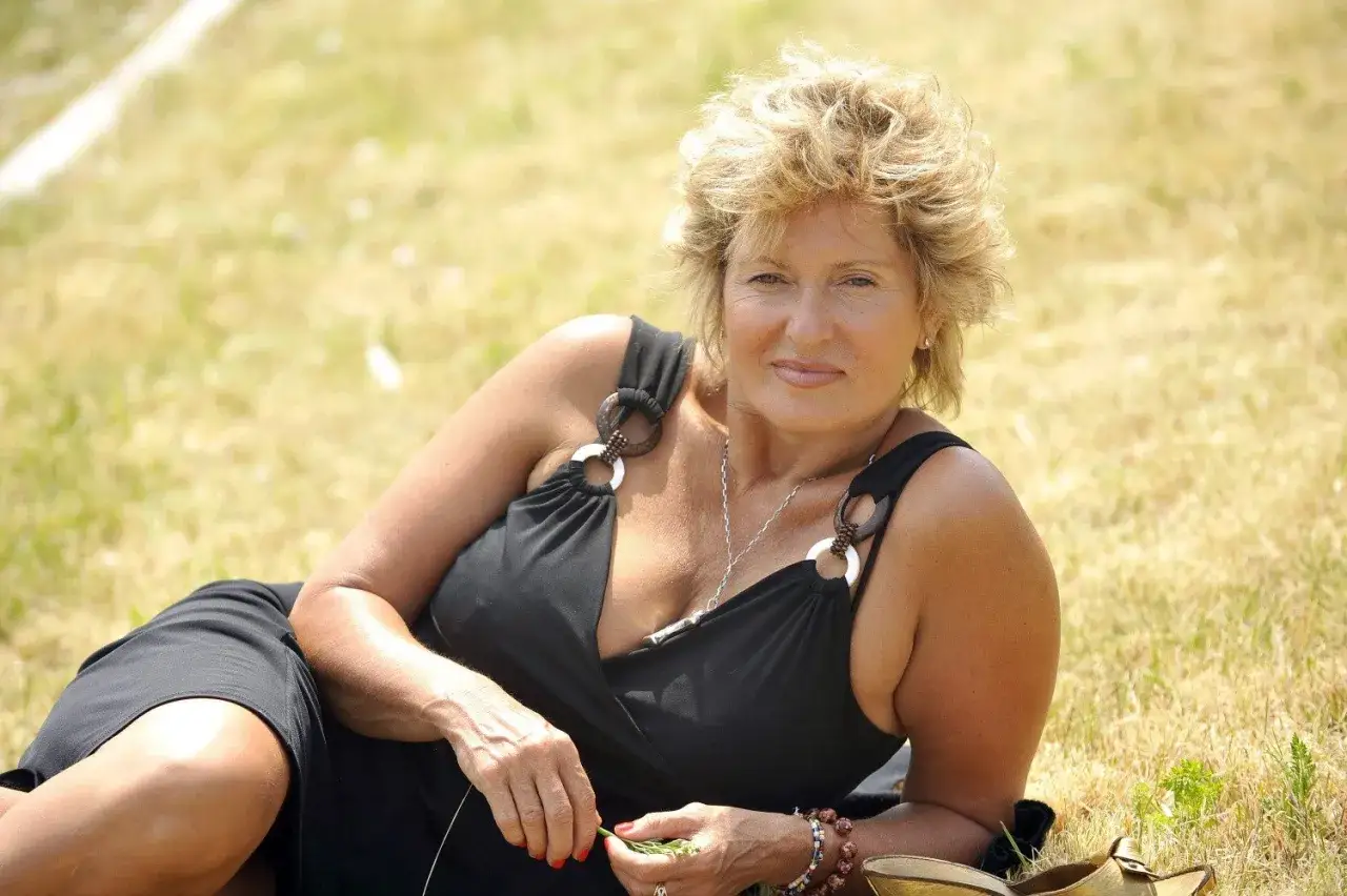 Ewa Kasprzyk w czarnej sukience, relaksująca się na trawie. Jej uśmiech mógłby pojawić się w niejednym z jej filmów.