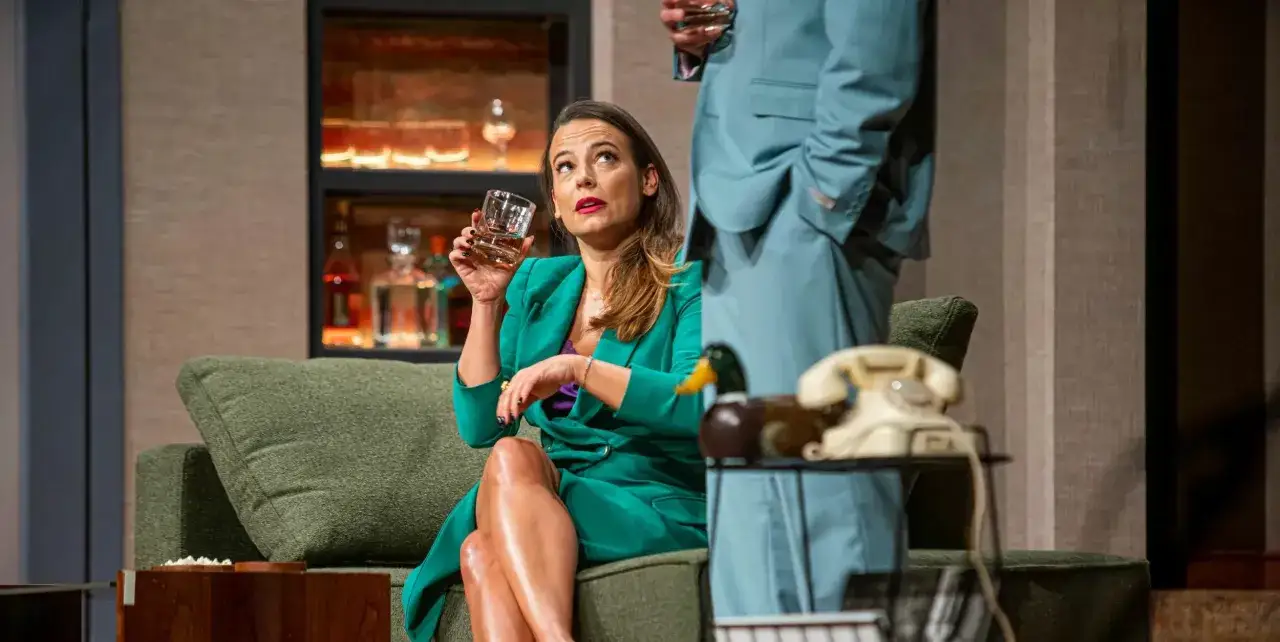 Kobieta w zielonej marynarce trzyma kieliszek, siedząc na kanapie. Scena z jednej z najlepszych komedii teatralnych.