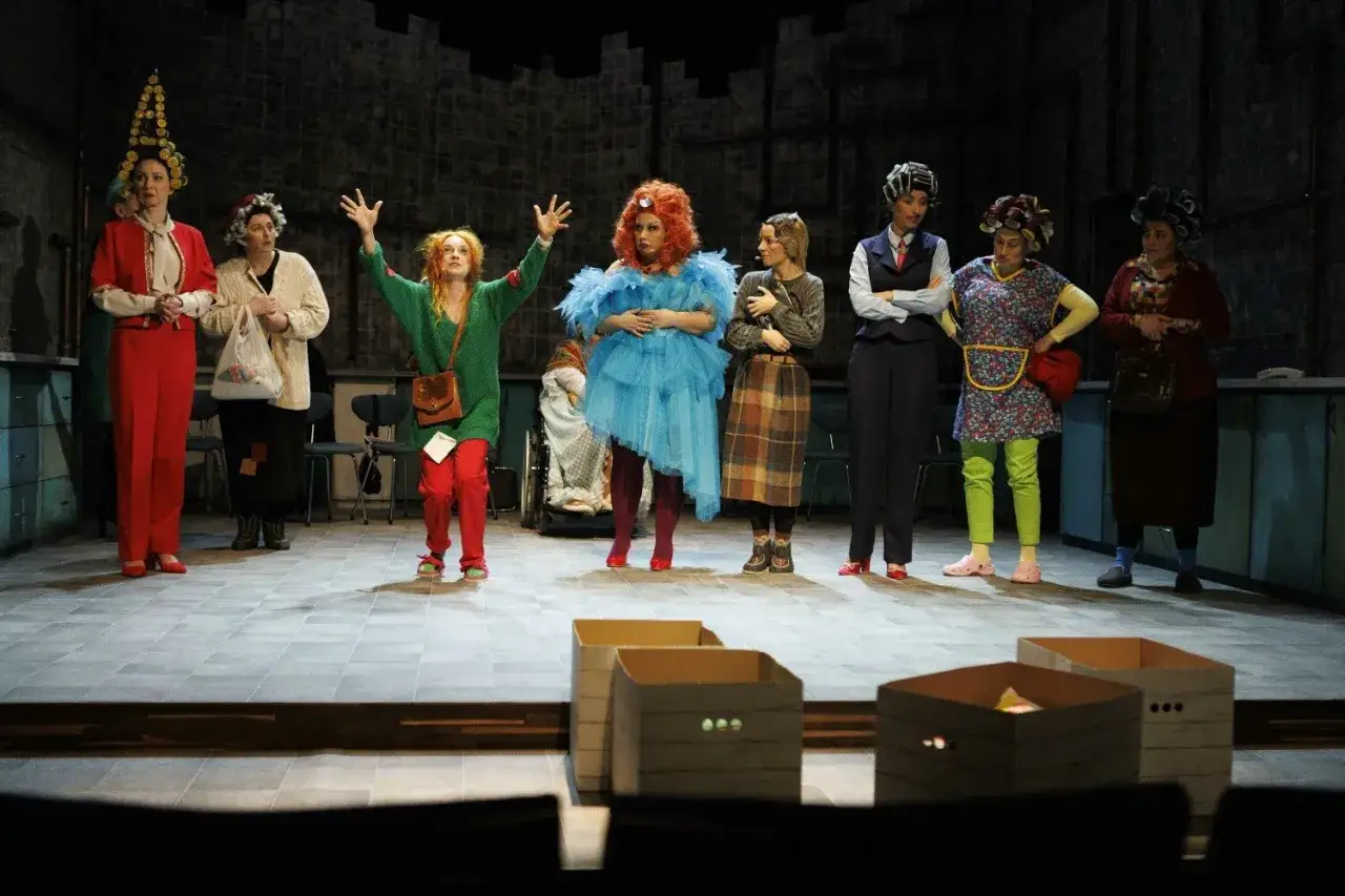Aktorzy na scenie Teatru Muzycznego Praga w barwnych strojach. Jedna z postaci w zielonym swetrze wznosi ręce.