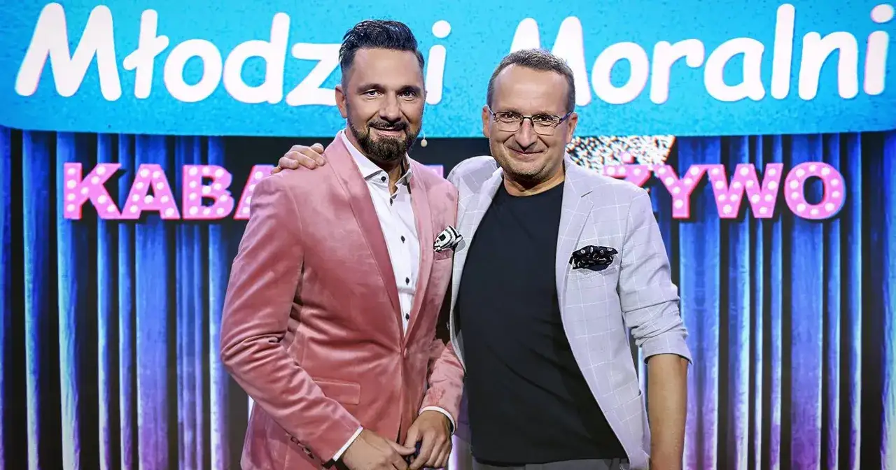 Dwóch mężczyzn na scenie, jeden w różowym aksamitnym garniturze, drugi w jasnej kraciastej marynarce. Tło z napisem