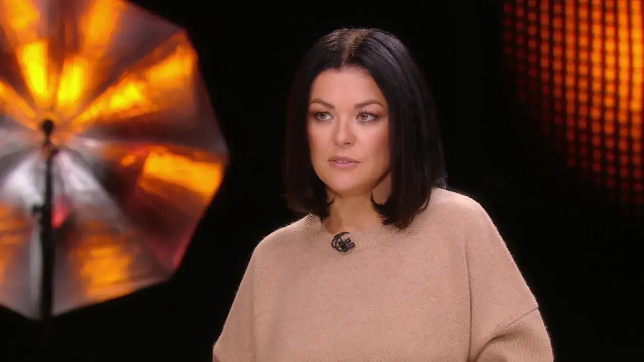 Katarzyna Cichopek w beżowym swetrze, w tle pomarańczowe światła. Może opowiada o swoich filmach?