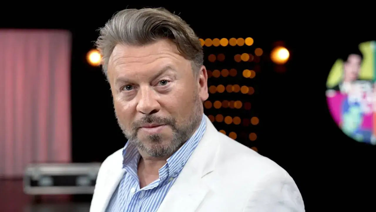 Michał Milowicz w białej marynarce, znany z filmów, seriali i programów, na tle świateł.