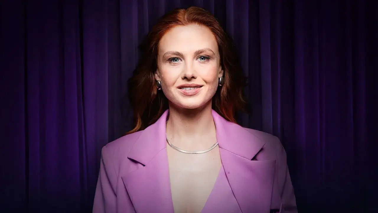 Anna Karczmarczyk w różowej marynarce, z rudymi włosami i niebieskimi oczami, uśmiecha się na tle fioletowej kurtyny. Jej filmy, seriale i programy to świetna rozrywka.